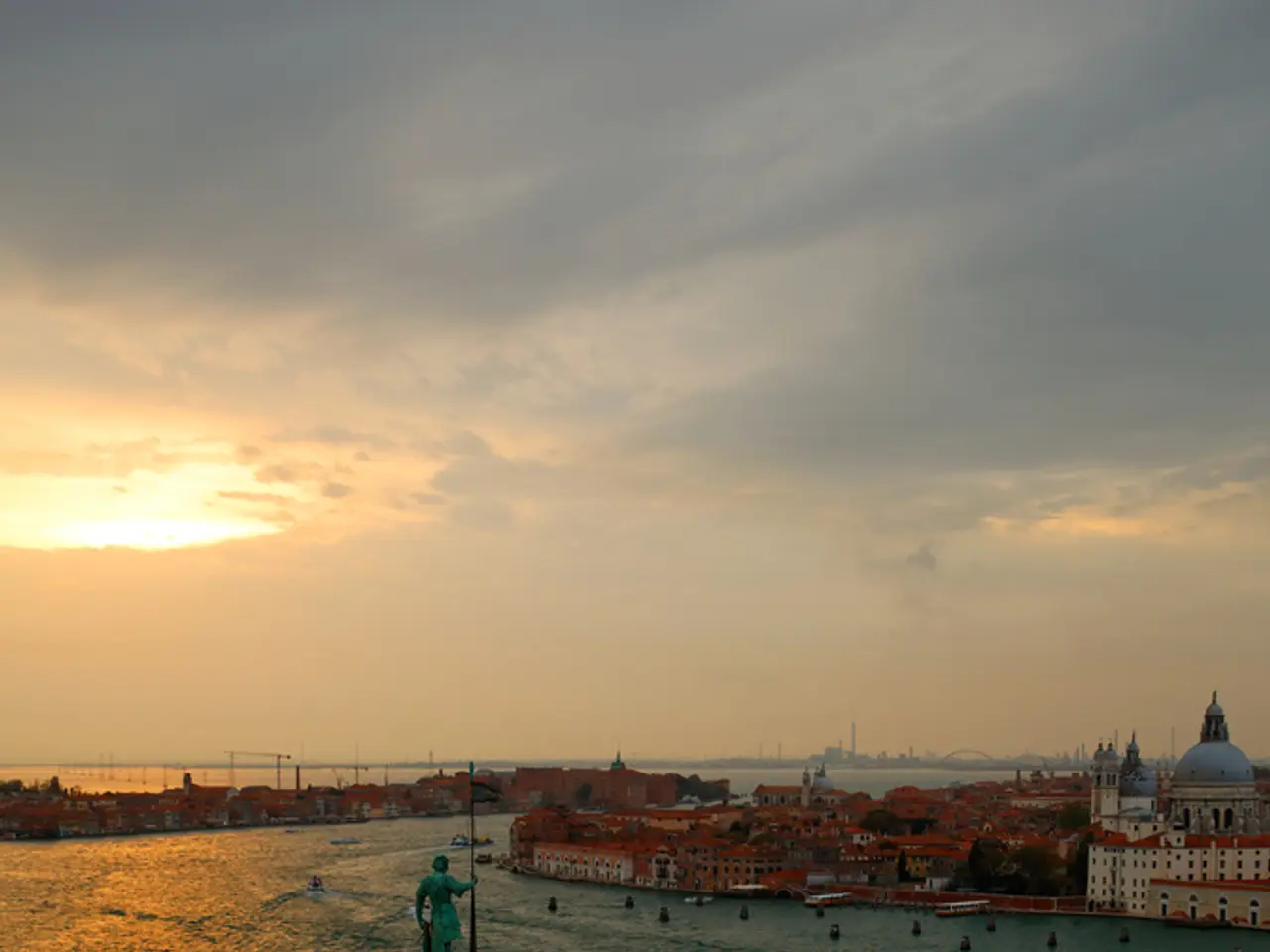 This picture shows few buildings and we see few boats in the water and a cloudy sky with sunlight.